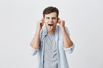 Emotional young male student in blue shirt grimacing, mocking, making faces, pulling ears, sticking out his tongue, looking at camera with crazy and stupid expression on his face.