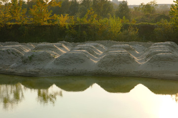 Sand dunes on the shore of the fishpond. A reflection of the dunes on the calm surface of the lake. The arrangement of sand dunes points to order. The water of the lake brings a balance of contrasts.