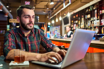 Portrait of modern young man working with laptop sitting  at bar counter in beer pub, copy space