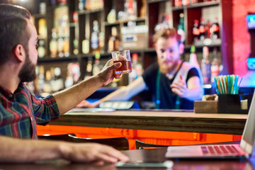 Portrait of modern young man toasting to bartender while working with laptop in pub, copy space