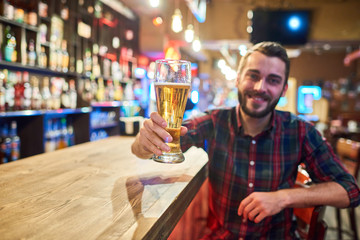 Portrait of smiling  bearded  man holding tall glass of beer cheering looking at camera while...