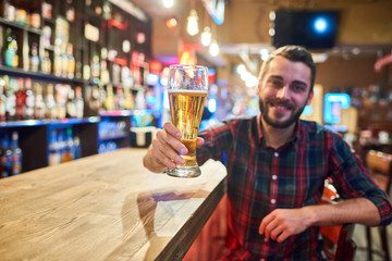 Portrait of smiling  bearded  man holding tall glass of beer cheering looking at camera while having fun in pub, copy space