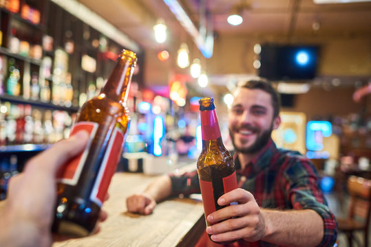 Portrait of happy bearded man clinking beer bottles with friends while having fun in pub sitting at bar counter, focus on foreground
