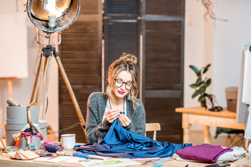 Young female fashion designer working with blue fabric sitting at the beautiful office with different tailoring tools on the table