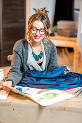 Young female fashion designer working with blue fabric sitting at the beautiful office with different tailoring tools on the table