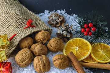 Winter abstract arrangement in studio on black background with snow, cinnamon sticks, lemon slices, frosty fir cone and braches and some small presents near a sack full of nuts.