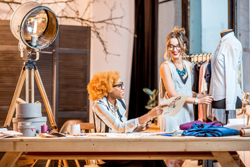 Two female multi-ethnic fashion designers working at the office with different tailoring tools and clothes
