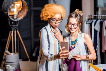 Two female multi-ethnic fashion designers working with smartphone standing at the office with different tailoring tools and clothes
