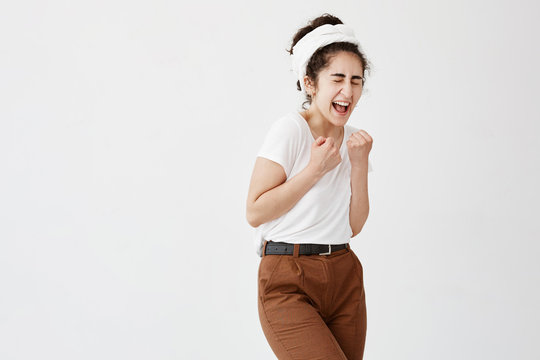 Lucky female with curly bushy hair in bun in white t-shirt keeping fists clenched opening her mouth and closing eyes with pleasure feeling great excitement and triumph isolated against white wall