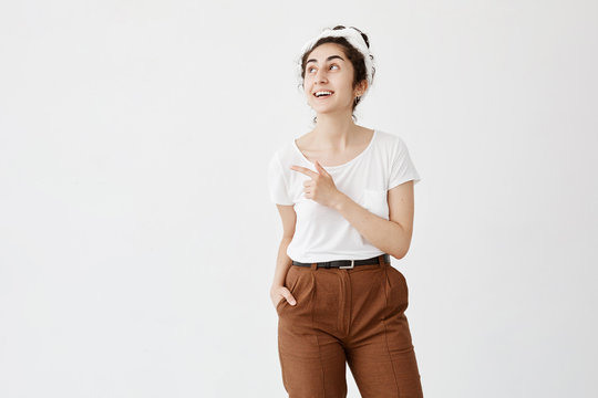 Smiling woman with dark wavy hair in bun in casual clothes posing against white studio wall pointing at copy space for your advertisment or text. Positive girl with hair bun advertising something