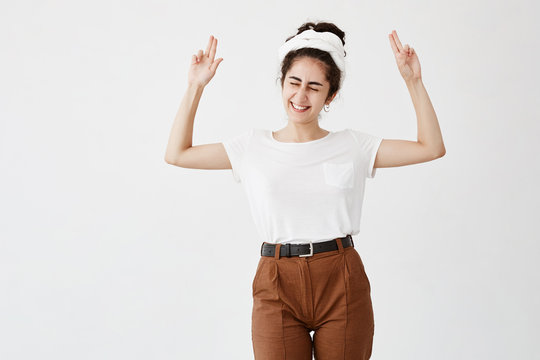 Excited overjoyed dark-haired woman points at copy space, closing her eyes, smiles broadly, advertisizes. Happy female in white t-shirt poses against white wall with copy area for promotional text