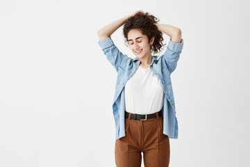 Youth and happiness concept. Brunette teenage girl in denim shirt and brown trousers, posing against white background with closed eyes, playing with her long wavy hair. Young woman smiles pleasantly