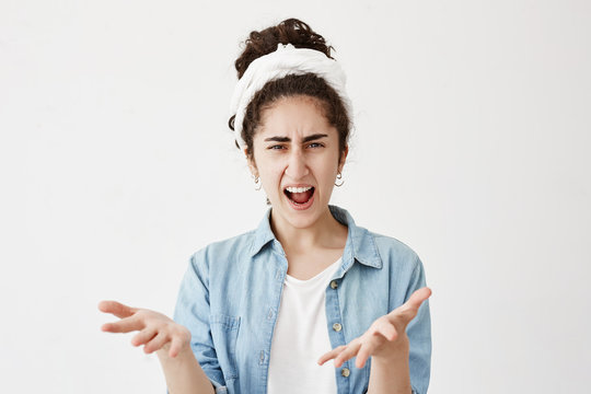 Close up isolated portrait of young dark-haired annoyed angry woman in do-rag and denim shirt holding hands in furious gesture. Young female in white T-shirt expresses negative emotions and feelings