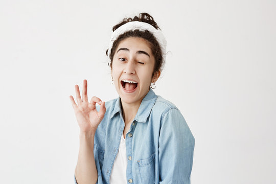 Positive girl with dark hair in bun wearing denim shirt and do-rag, blinking and opening mouth with joy showing ok sign being glad after meeting with his boyfriend isolated against white background.