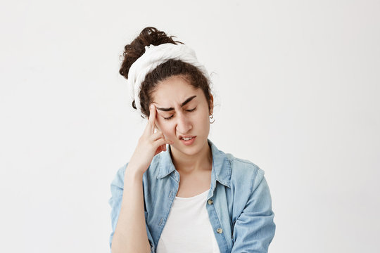 Picture of dark-haired pensive female model in white do-rag with closed eyes keeps hand on temple, suffers from headache after being worried, feels badly. Negative emotions and face expression.