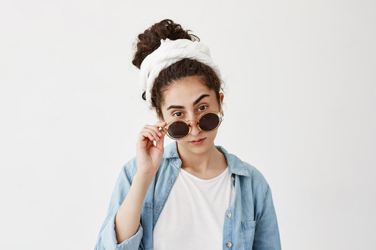 Pretentious dark-haired girl wearing white do-rag and sunglasses looks with appeal at camera, dressed in denim shirt, poses against white background. Face expression ad body language concept.