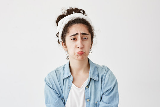 Indoor shot of dark-haired upset female being abused by someone, curves lips, frowns, looks unhappy, wears denim shirt, isolated against white background. Negative human emotions and feelings
