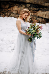 Fototapeta na wymiar Beautiful woman stands on the road in snowy mountains in grey wedding dress with bouquet in hands