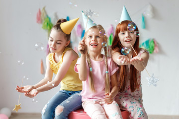 Portrait of three beautiful girls wear festive caps, play with bubbles, sit together on chair,...