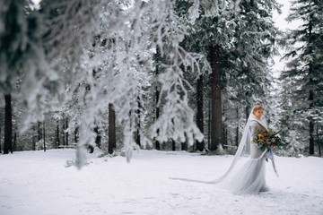 Beautiful woman stands on the snowy pine forest in grey wedding dress with bouquet in hands and long veil