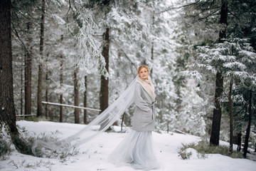 Beautiful woman stands on the snowy pine forest in grey wedding dress with bouquet in hands and long veil