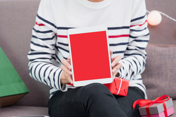 Close up woman hand holding tablet sitting on sofa with gift and present box in Christmas and new year party,winter festive holiday celebration,Mock up red screen for display or advertise promotion.