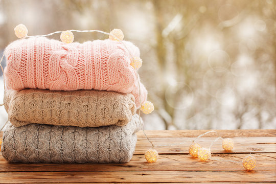 Cozy pile of knitted sweaters on wooden table on winter nature background with snow