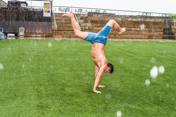 Man Playing in Rain, foggy, wet, grainy feel. Shirtless, half naked, wearing blue shorts, barefoot, arms supporting on ground, a young guy flipping whole body upside down on air, in hot summer