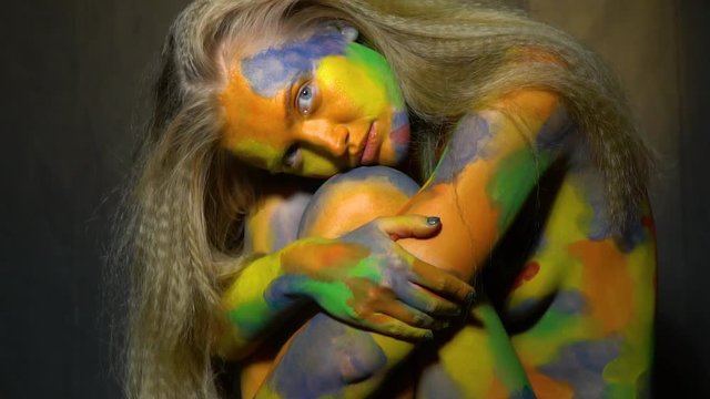 Mysterious long-haired blonde in multi-colored body art sitting with her knees bent