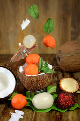 Tropical fruits: longan, rambutan and papaya ball-shaped pieces are falling into the coconut cup and around it. Ingredients for a cocktail or a dessert