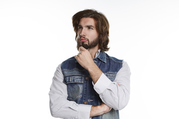 Picture of fashionable uncertain doubtful young bearded male in stylish clothes touching beard and looking sideways with pensive expression, feeling hesitant about making important decision