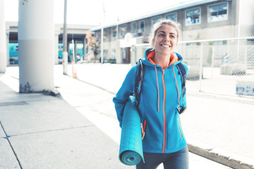 Obraz premium Athlete woman with backpack, headphones, yoga mat going to sport club on the city street. Smiling. Sport tight clothes.