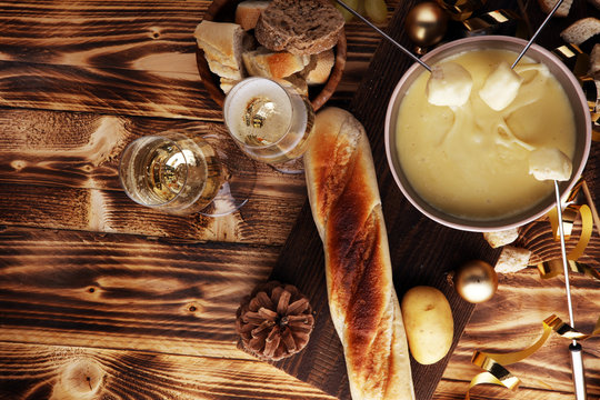 Gourmet Swiss fondue dinner on a winter evening with assorted cheeses on a board alongside a heated pot of cheese fondue with two forks dipping bread and white wine behind in a tavern or restaurant.