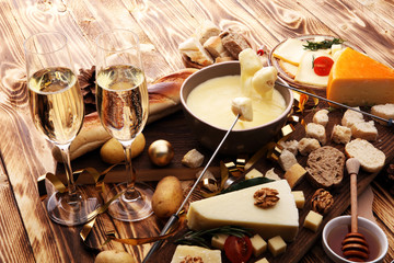 Gourmet Swiss fondue dinner on a winter evening with assorted cheeses on a board alongside a heated pot of cheese fondue with two forks dipping bread and white wine behind in a tavern or restaurant.