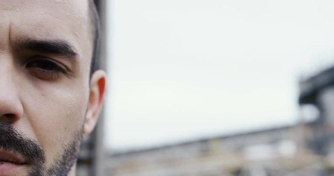 Close up of the half face of the handsome man with a black beard looking seriousy in the camera outdoor. Blurred plant on the background. Grey sky. Portrait