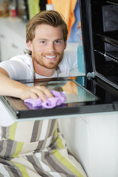 happy young man in overall cleaning oven with rag