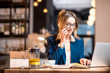 Young businesswoman strictly dressed in the suit talking phone while working with laptop at the modern cafe interior