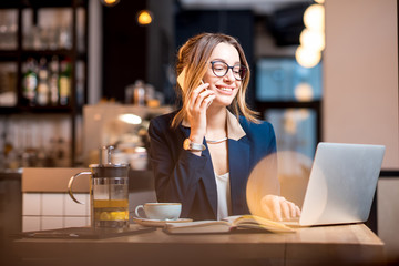 Young businesswoman strictly dressed in the suit talking phone while working with laptop at the modern cafe interior