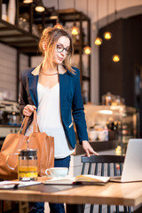 Young businesswoman strictly dressed in the suit coming with bag at the cafe table having a coffee break