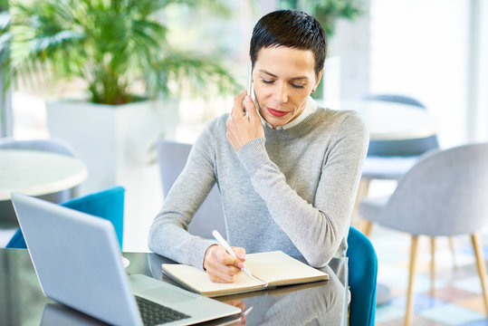 Portrait of successful modern businesswoman speaking by phone and making notes while working in cafe during lunch break