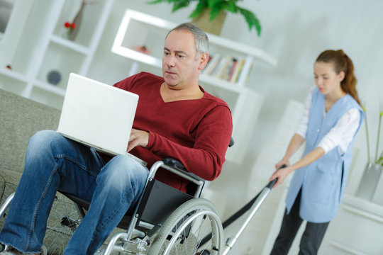 disabled man at home using laptopwoman cleaning in background