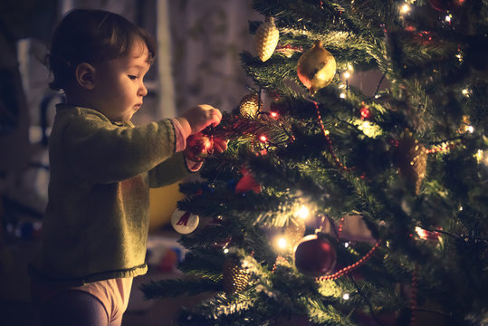 Baby is hanging decoration on Christmas tree, child expects New Year at home