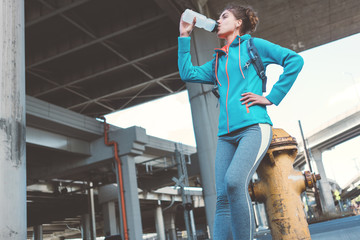 Athlete young girl full of strength and self-confidence with backpack, headphones resting after workout on the city street. Drinking water. Sport tight clothes.
