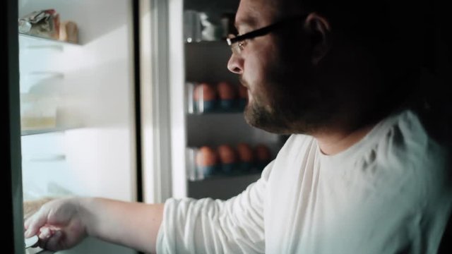 Fat man opens a refrigerator at night and eats a piece of cake, night gluttony and unhealthy food