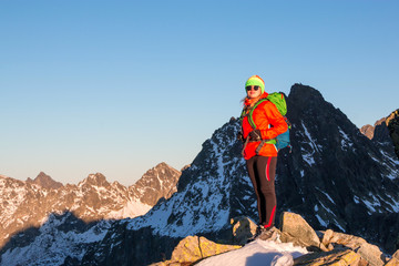 Smiling attractive young woman hiking in winter high mountains with backpack, she looking at the peak of mountain at sunset.