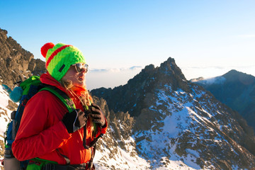 Smiling attractive young woman hiking in winter high mountains with backpack, she looking at the peak of mountain at sunset.