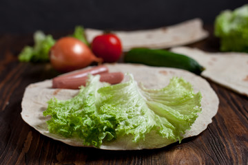 cooking burrito with greens, cucumbers, tomatoes and sausages on a round wooden board on a wooden background