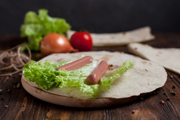 cooking burrito with greens, cucumbers, tomatoes and sausages on a round wooden board on a wooden background