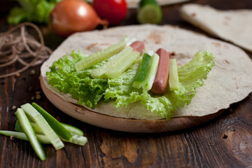 cooking burrito with greens, cucumbers, tomatoes and sausages on a round wooden board on a wooden background