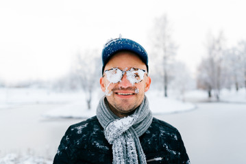 Fototapeta na wymiar Closeup portrait of male face covered with snow. Crazy cheerful funny comic emotions. Odd bizarre unusual winter idea. Stylish boy wear glasses, hat, scarf. Unique person. Cryotherapy. Frozen skin